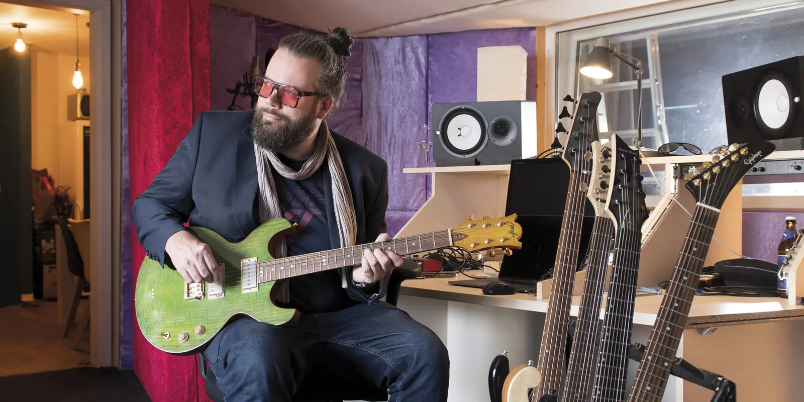 Tobias Langer sitzt mit einer grünen E-Gitarre in der Hand auf einem Barhocker, im Vordergrund sind weitere E-Gitarren auf einem Ständer aufgestellt.