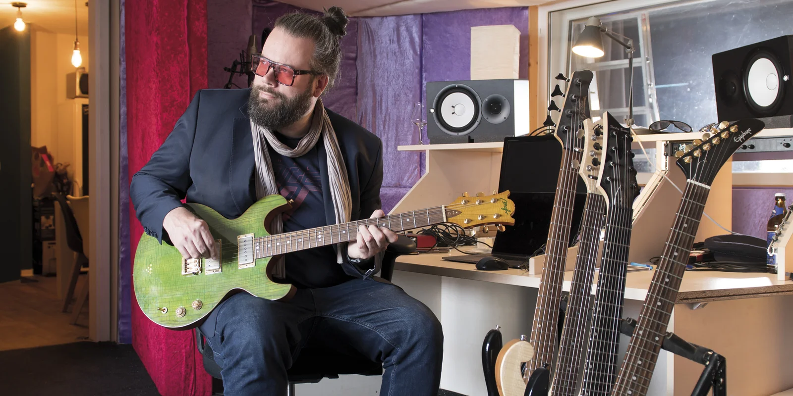 Tobias Langer sitzt mit einer grünen E-Gitarre in der Hand auf einem Barhocker, im Vordergrund sind weitere E-Gitarren auf einem Ständer aufgestellt.