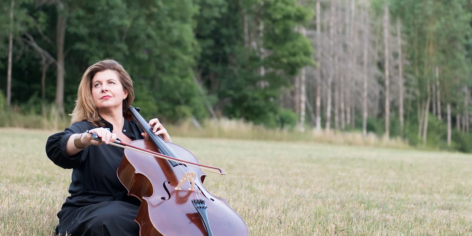 Lucile Chaubard auf einer Wiese sitzend Cello, im Hintergrund ein Nadelwald.