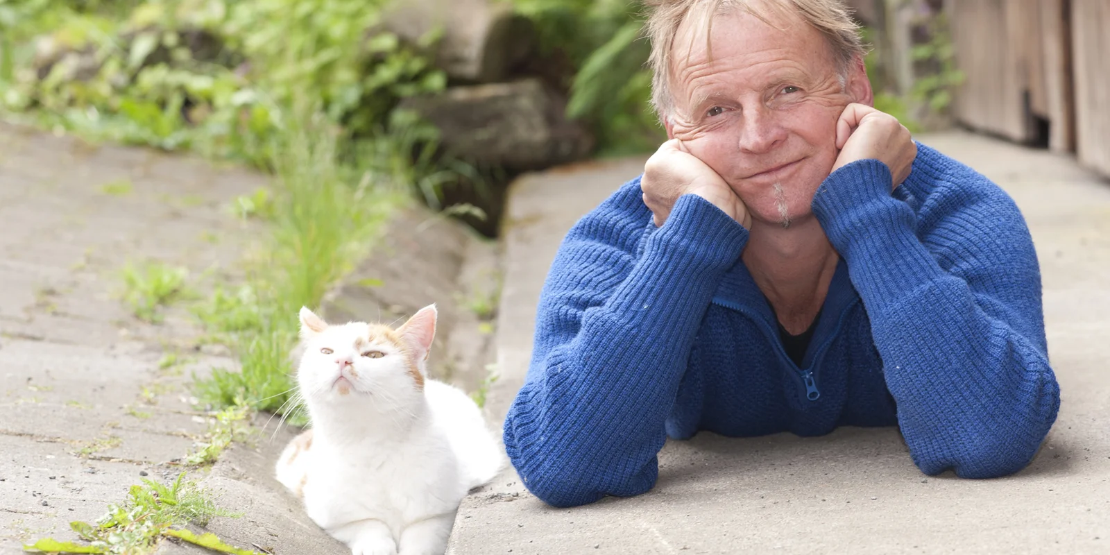 Nik J. Lucht liegt bäuchlings neben einer weißen Katze auf einem Betonboden und Blickt in die Kamera. Den Kopf hat er in die Hände gestützt.