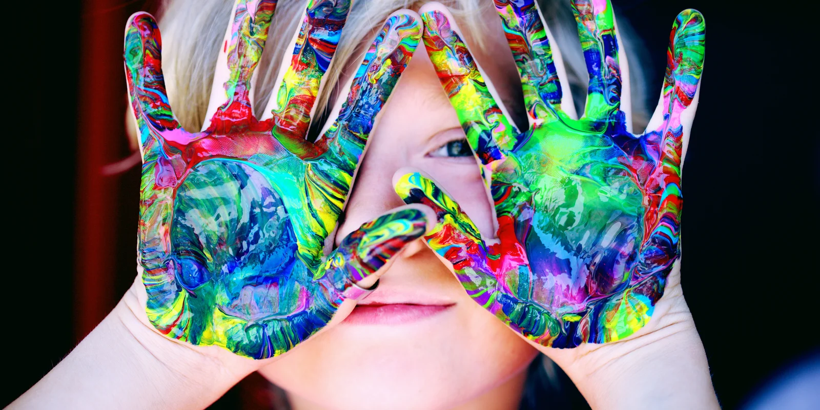 Ein Kind hält sich mit bunter Farbe bemalte Hände vor sein Gesicht und blickt hindurch.