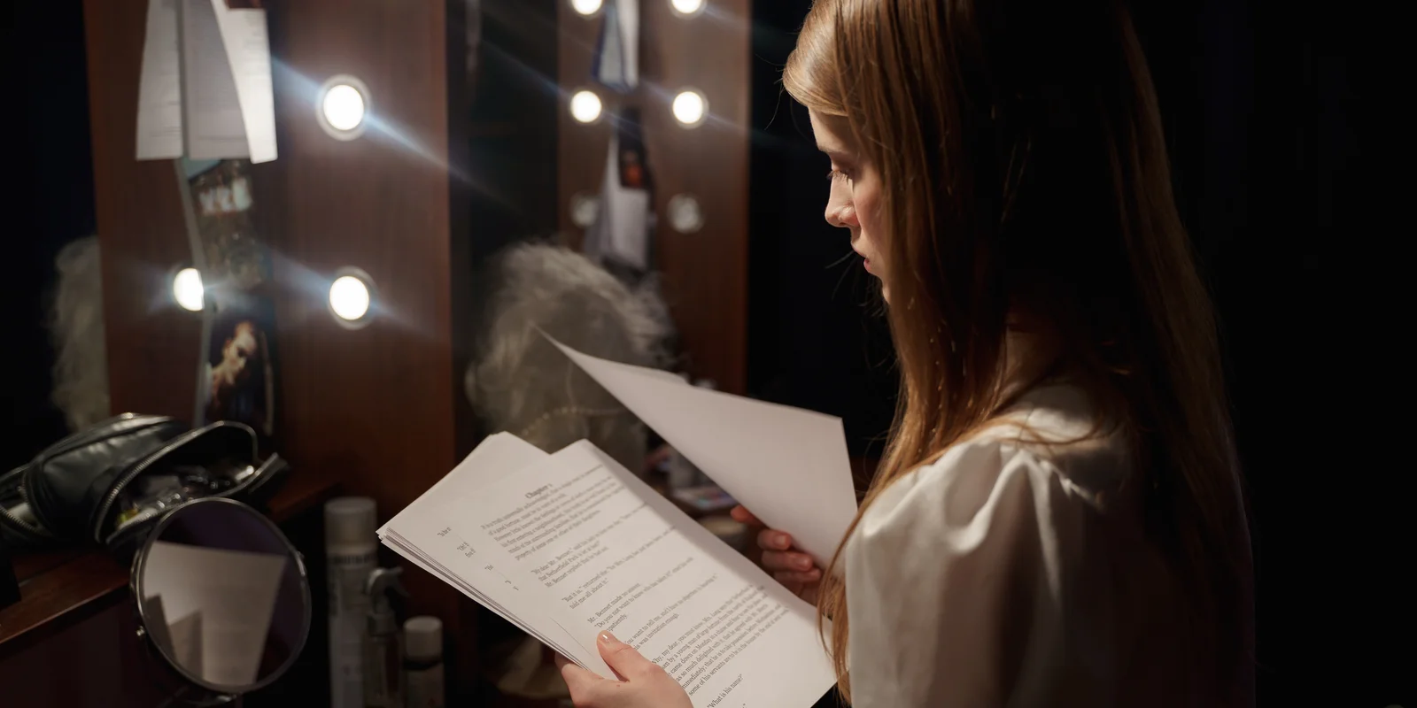 Eine junge Frau steht vor einem beleuchteten Spiegel, in den Händen hält sie mehrere Textblätter.