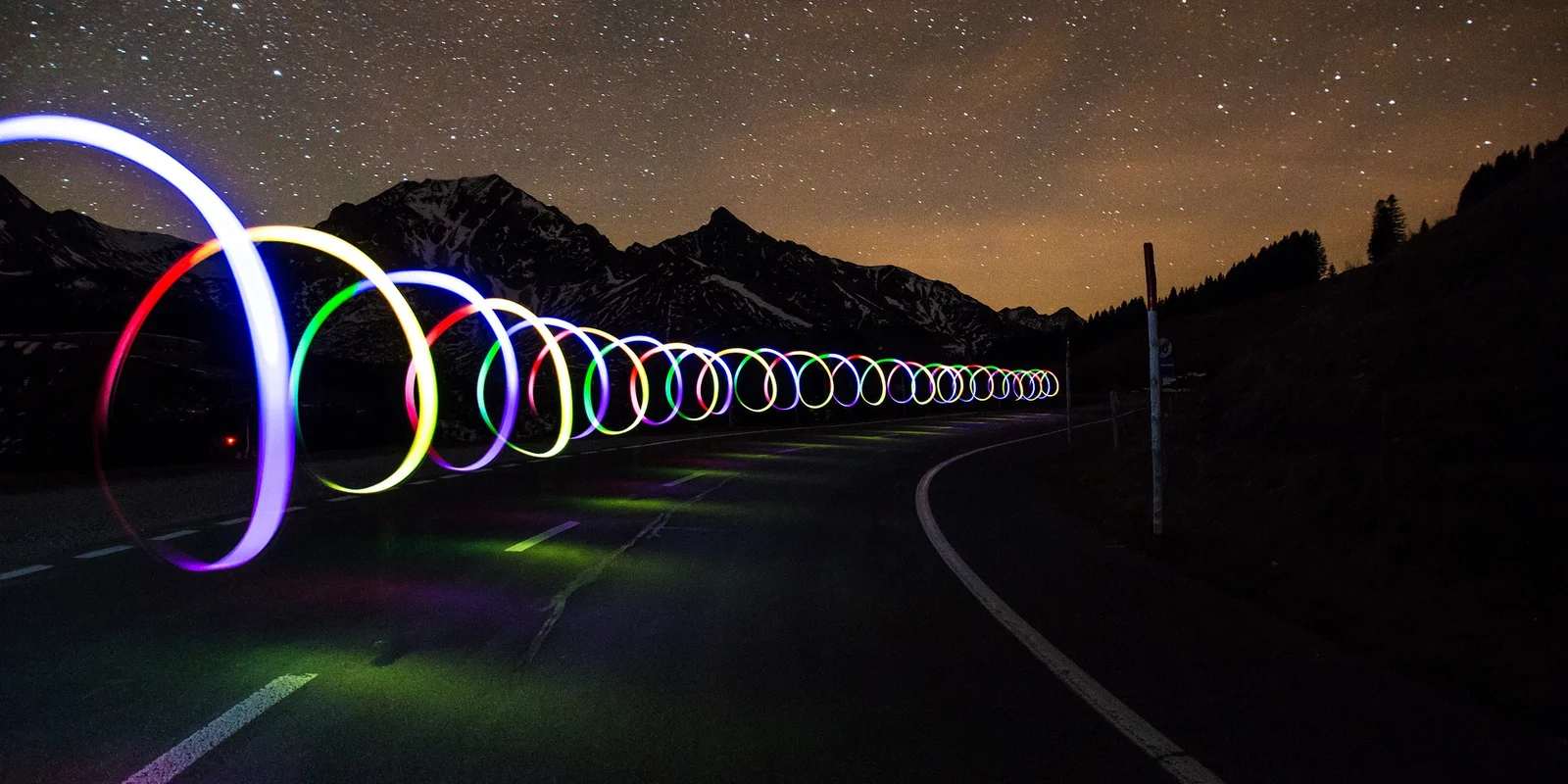 Eine Straße bei nacht mit Sternenhimmel. Über der Straße "schweben" Lichtzeichnungen: eine spiralförmige farbige Lichtröhre
