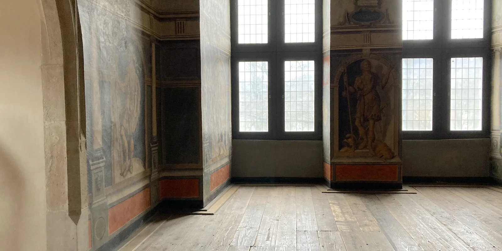 Blick in einen Raum mit bunten Wandfresken und hohen geschnitzten Holzdecken, erhellt von zwei raumhohen Fenstern