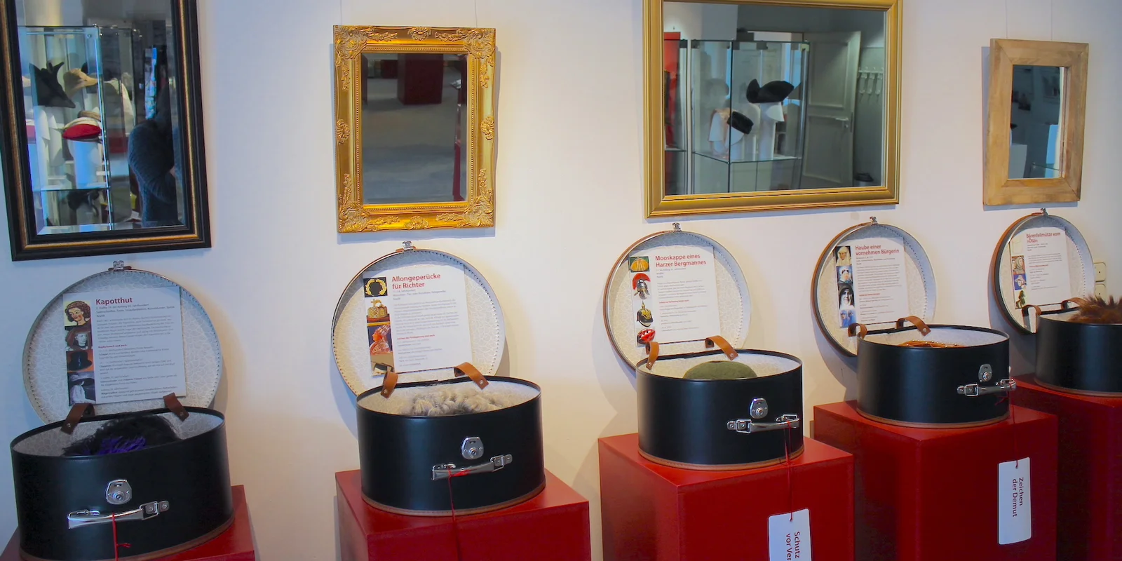 An einer Wand, an der verschiedene Spiegel hängen, stehen roten Podest mit offenen Hutschachteln, in denen unterschiedliche Hüte und Erläuterungstexte zu sehen sind