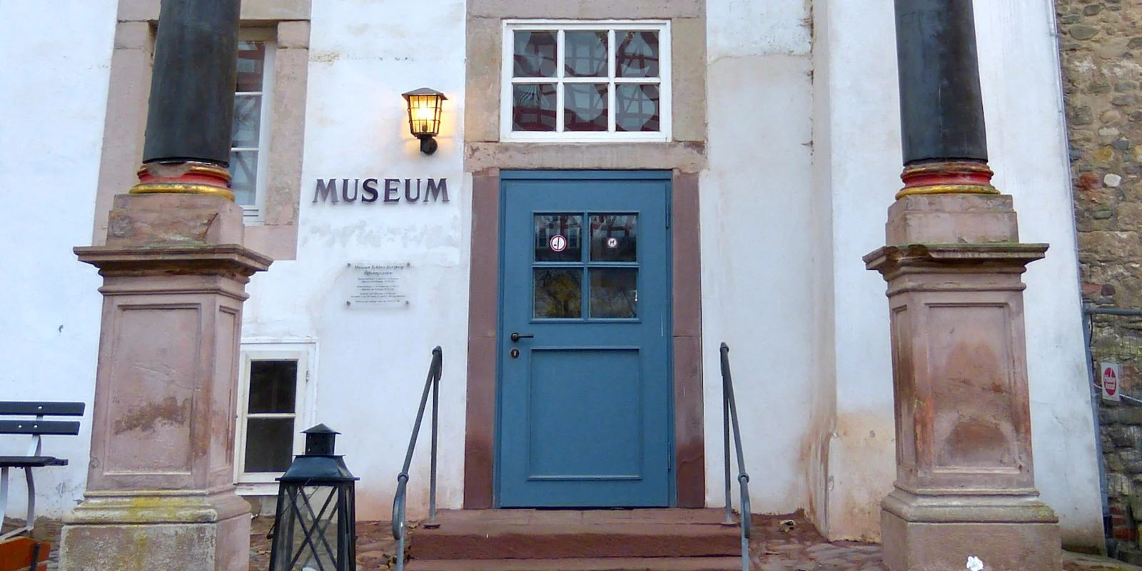 Einige Stufen führen zwischen zwei Säulen hindurch zu einer blauen Tür, neben der das Wort Museum an der Wand steht