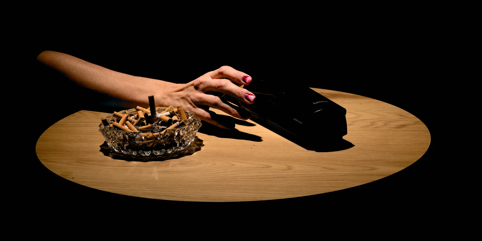 Ein Tisch, darauf ein voller Aschenbecher, von der linken Seite greift eine Hand nach einem Telefonhörer.