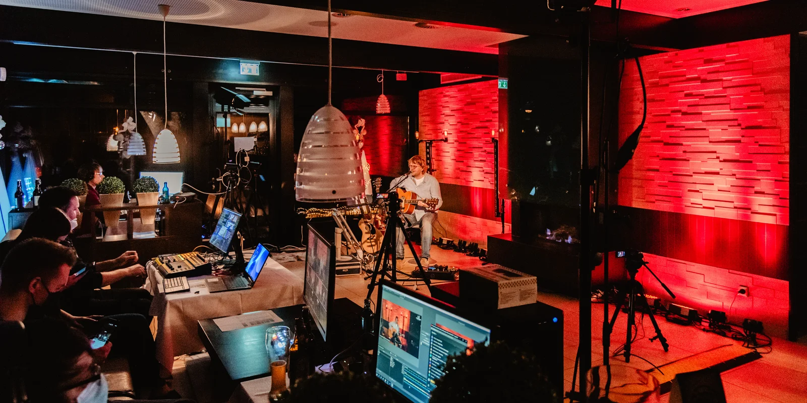 Ein blonder Gitarrist sitzt auf einem Barhocker vor einer rot beleuchteten Wand. Im Vordergrund sind mehrere Bildschirme aufgebaut, an denen Männer mit Corona-Masken sitzen, um einen Videostream zu koordinieren.