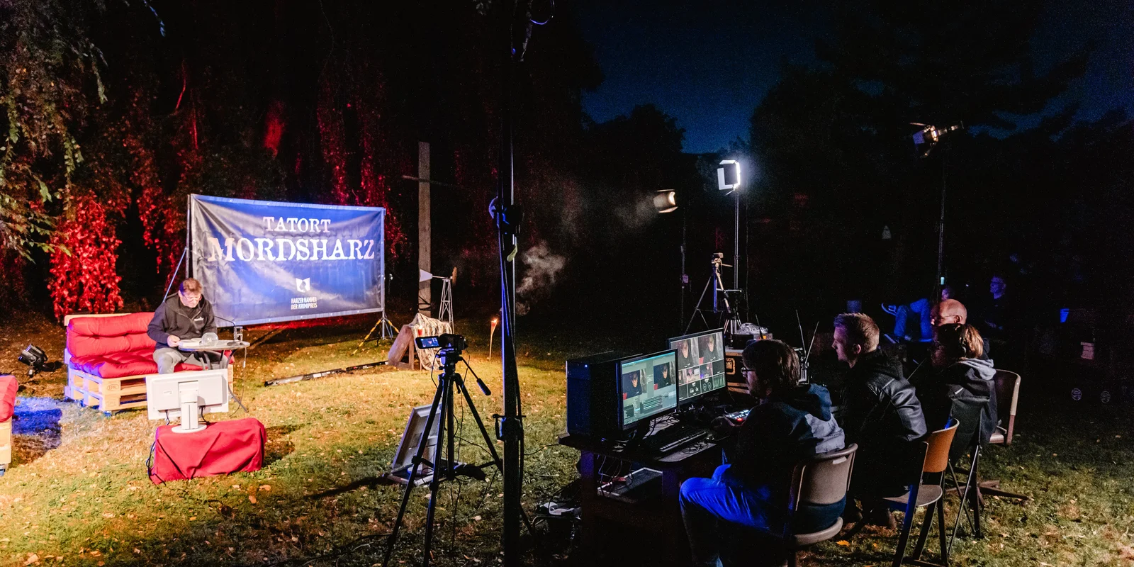 Ein Mann sitzt draußen vor Bäumen auf einem Palettensofa neben einem Plakat mit der Aufschrift: "Tatort Mordsharz". Davor sitzen 4 Personen vor Bildschirmen, Kamera und Scheinwerfern.