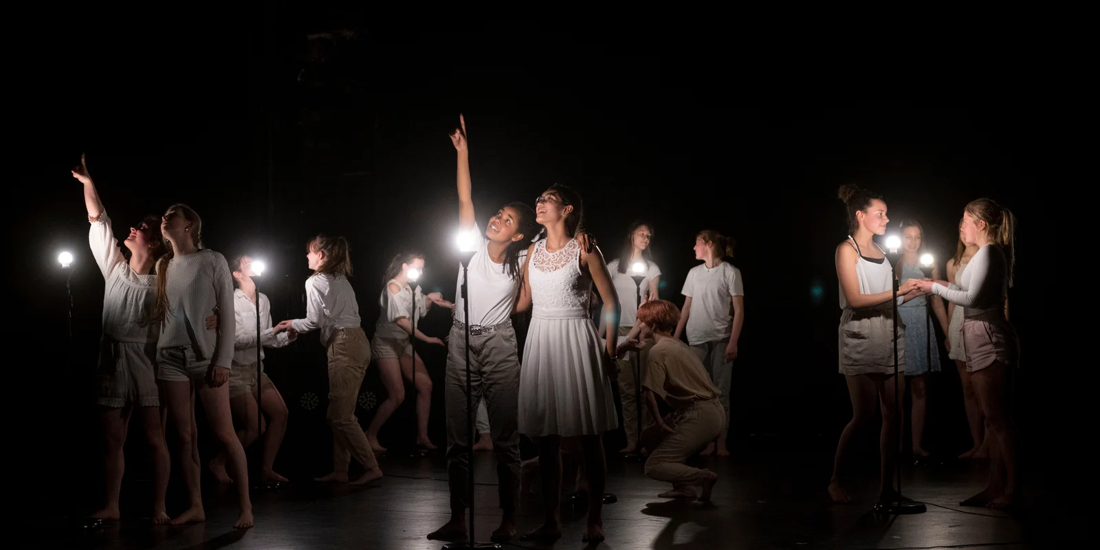 Auf einer abgedunkelten Bühne leuchten kleine weiße Lampen auf Ständern. Schülerinnen des Otto Hahn Gymnasiums sind in Zweierpärchen mit weißen T-Shirt und beigen Hosen oder weisen Kleidern zu sehen. Sie gucken sich gegenseitig in die Augen und halten sich an den Händen. zwei Schülerinnen zeigen mit dem Finger nach oben