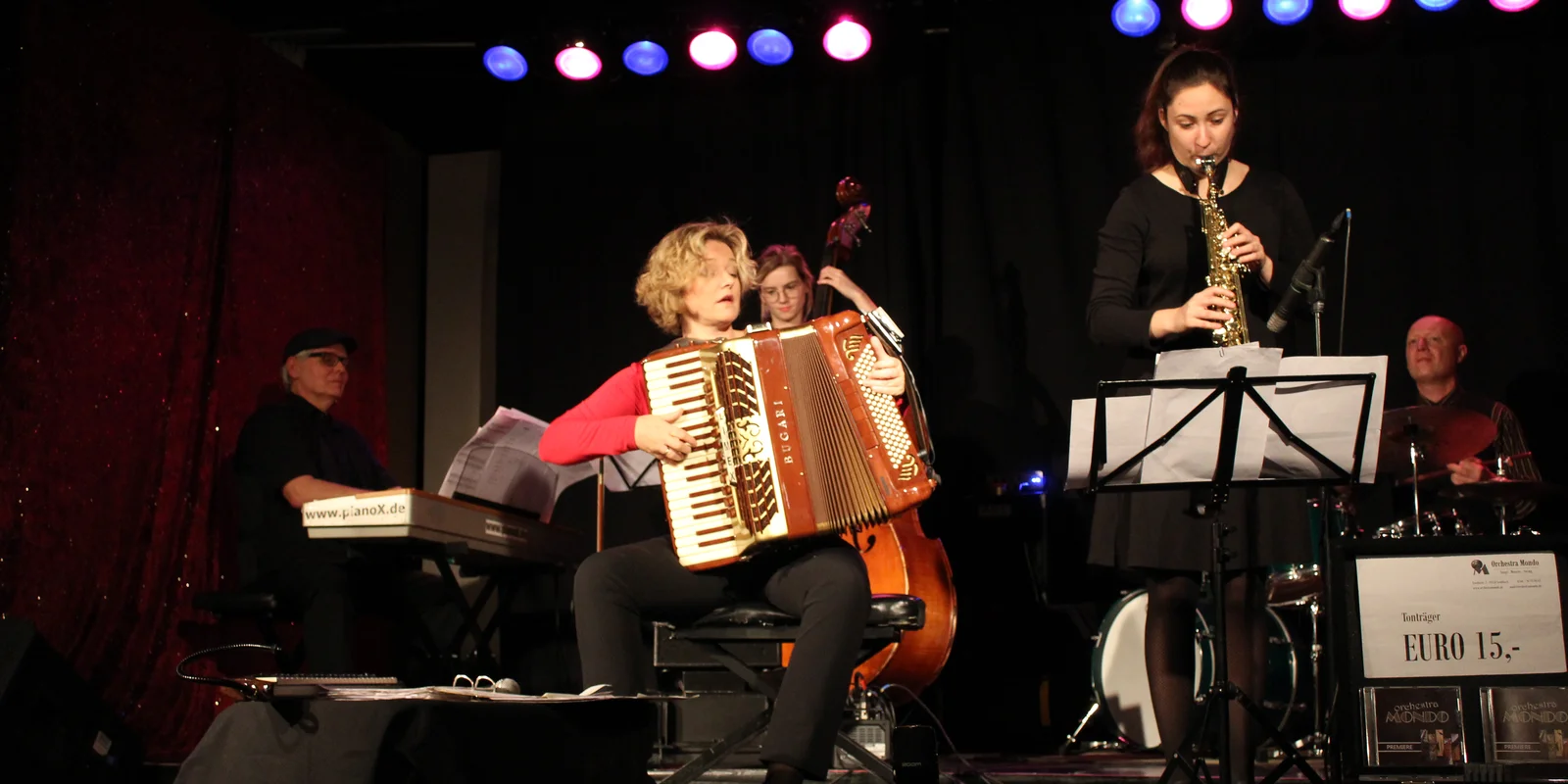 Eine blonde Frau mit rotem T-Shirt und schwarzer Hose sitzt auf einem Stuhl und spielt ein rotes Akkordeon. Im Hintergrund ist ein Pianist an einem elektrischen Keyboard, eine Contrabassspielerin, ein Schlagzeuger und neben ihr eine Saxophonspielerin