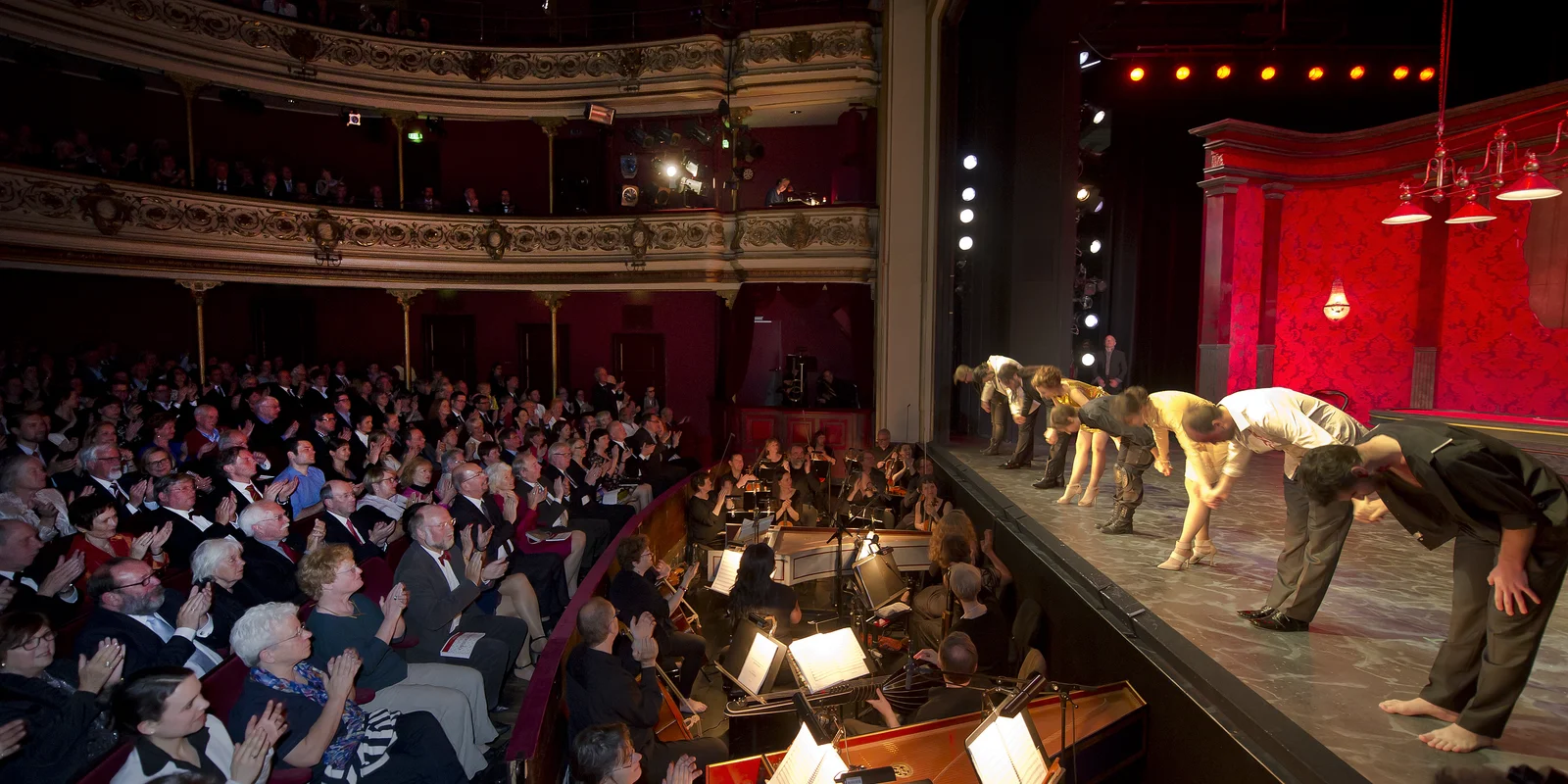 In einem klassischen Theater verbeugen sich Darstellerinnen und Darsteller auf der Bühne, im Orchestergraben klatschen Musikerinnen und Musiker und das Publikum applaudiert.