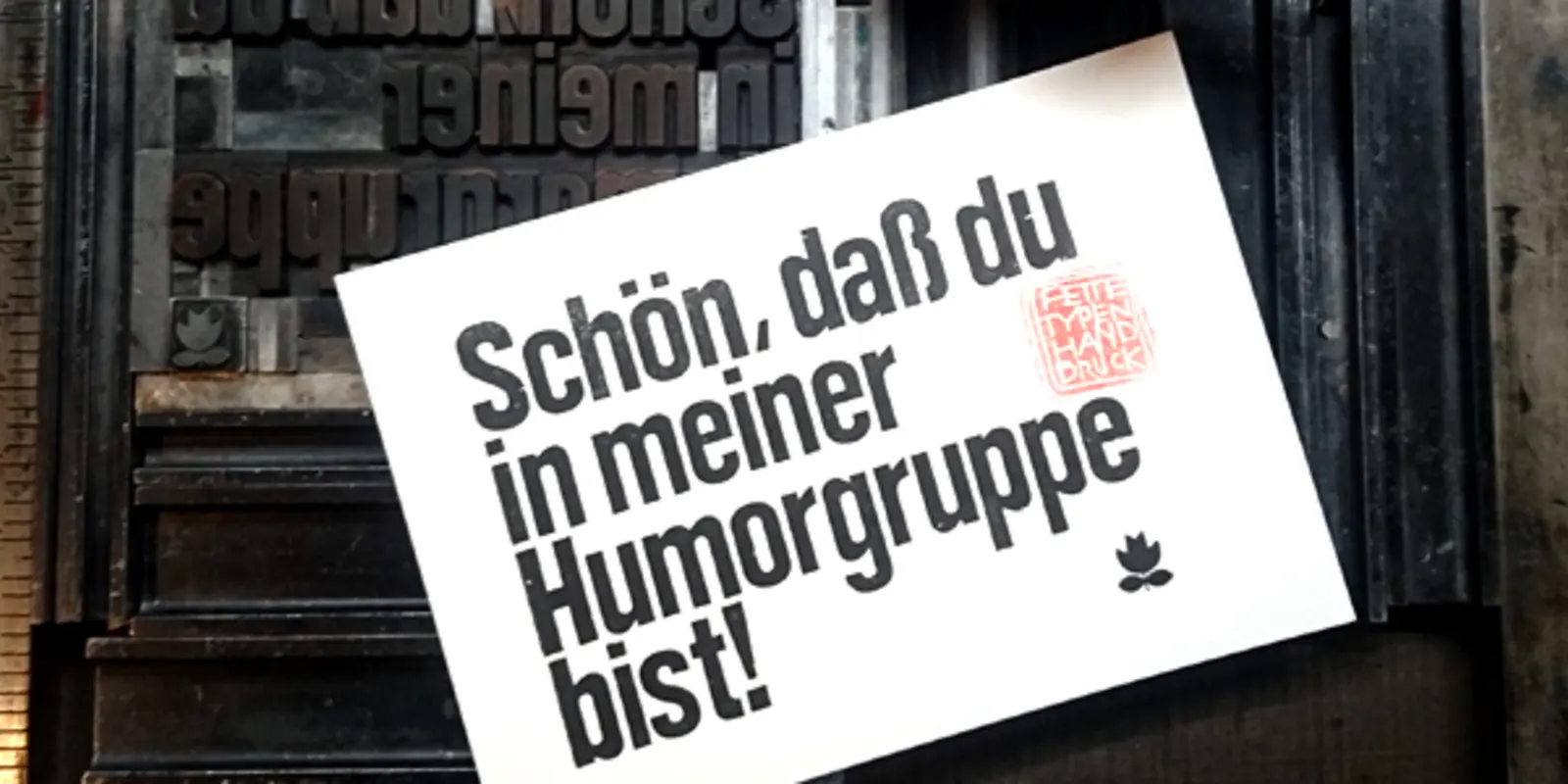 eine schwarze Druckmaschine mit Rolle. Auf einer weißen Karte steht in schwarzer Tinte gedruckt: "Schön, daß du in meiner Humorgruppe bist!"