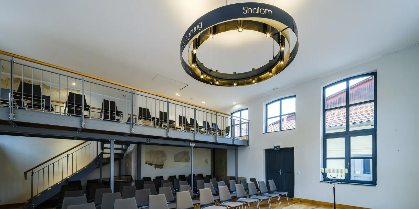 Der Raum in der Synagoge mit der Empore, schwarzen Stühlen und einem Kronleuchter mit drei schwarzen Stahlringen, in den jüdische Symbole eingearbeitet sind, An den weißen Wänden ist ein großes Fenster mit 8 Kacheln und zwei kleine Fenster.