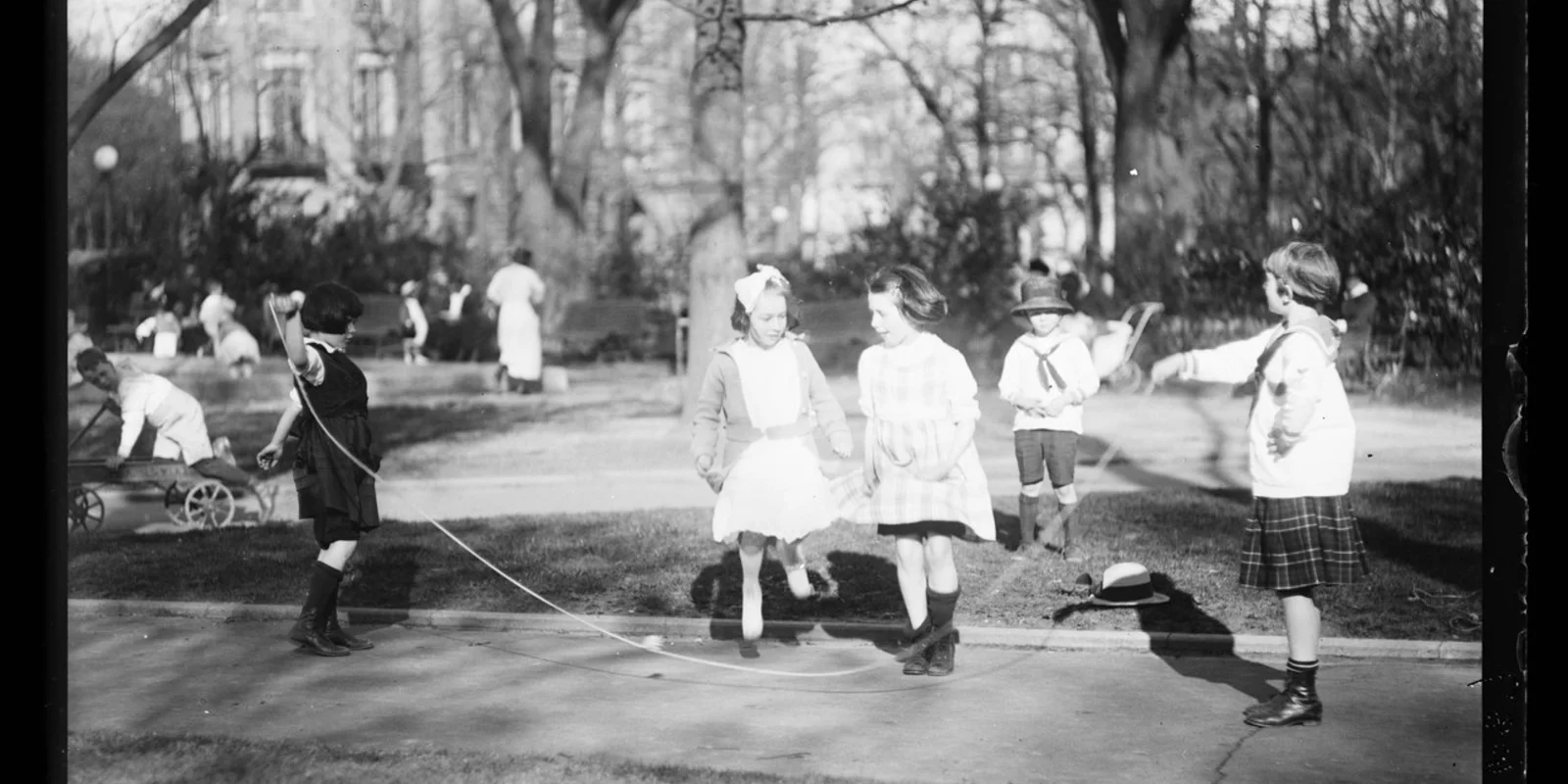 Schwarz-weiß Aufnahme von spielenden Kindern in einem Park im Jahr 1919. Im Vordergrund schwingen zwei Mädchen ein Seil, über das zwei weitere Mädchen springen.