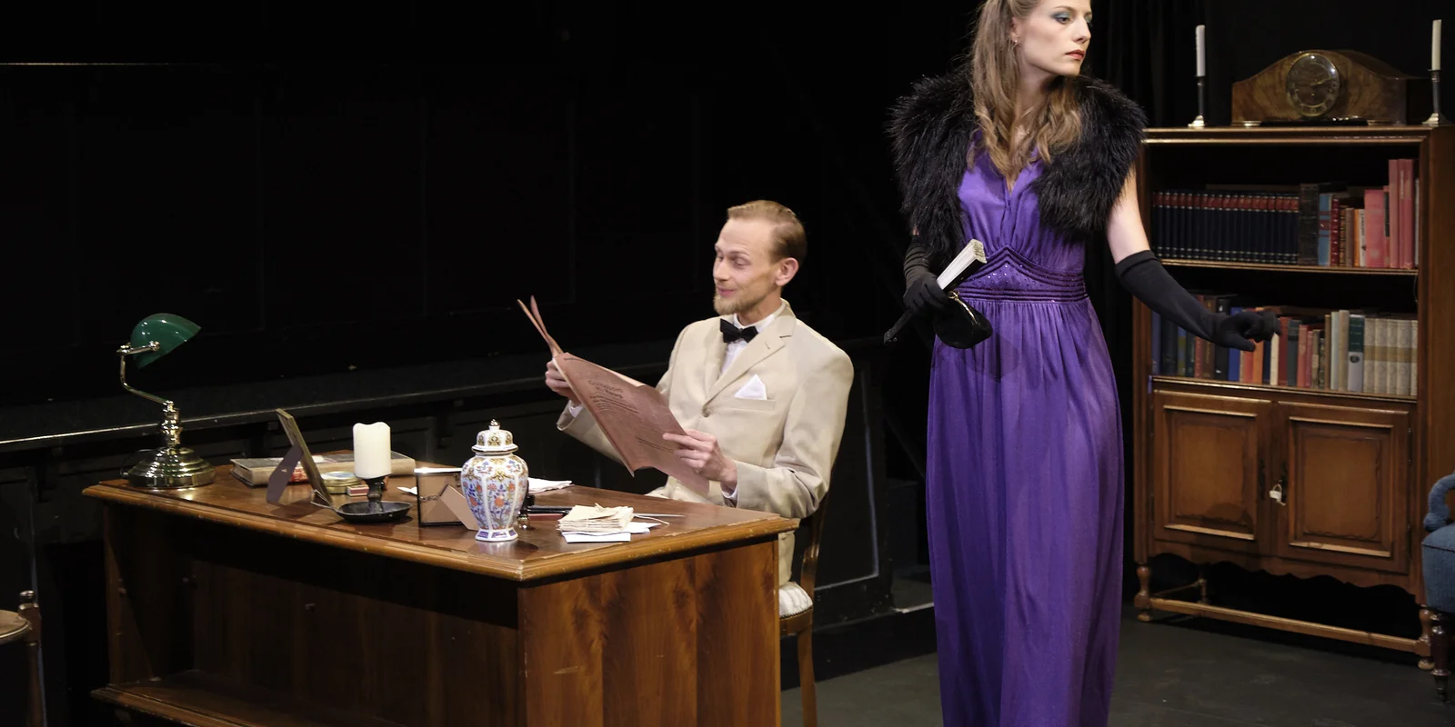 Foto. Bühne des ThOP. Ein Mann im Anzug sitzt an einem großen Holzschreibtisch. Eine elegant angezogene FRau (lila Kleid, Abendhandschuhe, Pelzstola) steht daneben 