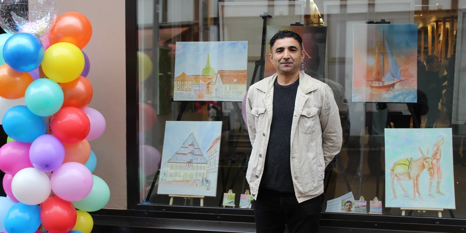 Der Künstler Abdulkarim Alhasan steht vor einem Fenster mit Bildern. Neben ihm sind bunte Luftballons