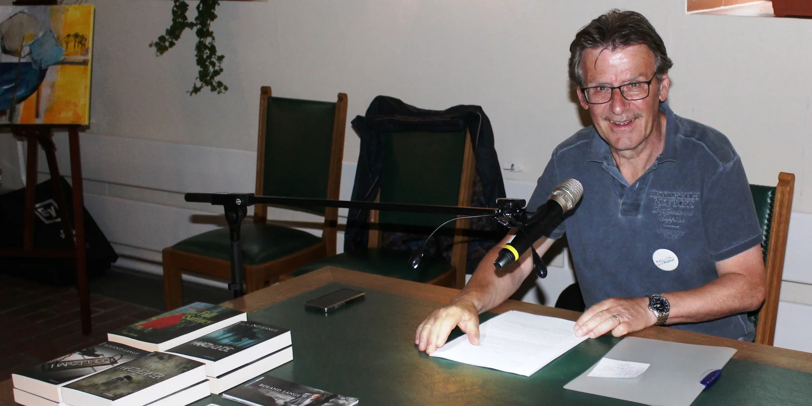 Roland Lange sitzt lächelnd an einem Schreibtisch, vor ihm ein aufgeschlagenes Buch und ein Mikrofon.
