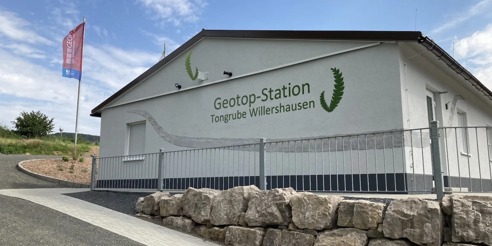 Ein weiß gestrichenes einstöckiges Gebäude, auf dessen Fassade in grüner Schrift "Geotop-Station Tongrube Willershausen" zu lesen ist