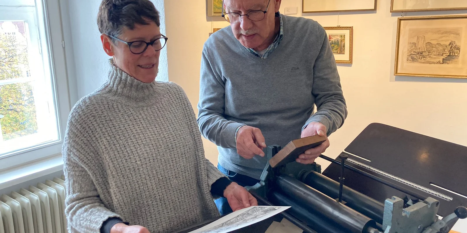 Barbara Brübach und Hermann Staub stehen vor einer Andruckpresse und halten ein bedrucktes Papier in der Hand 