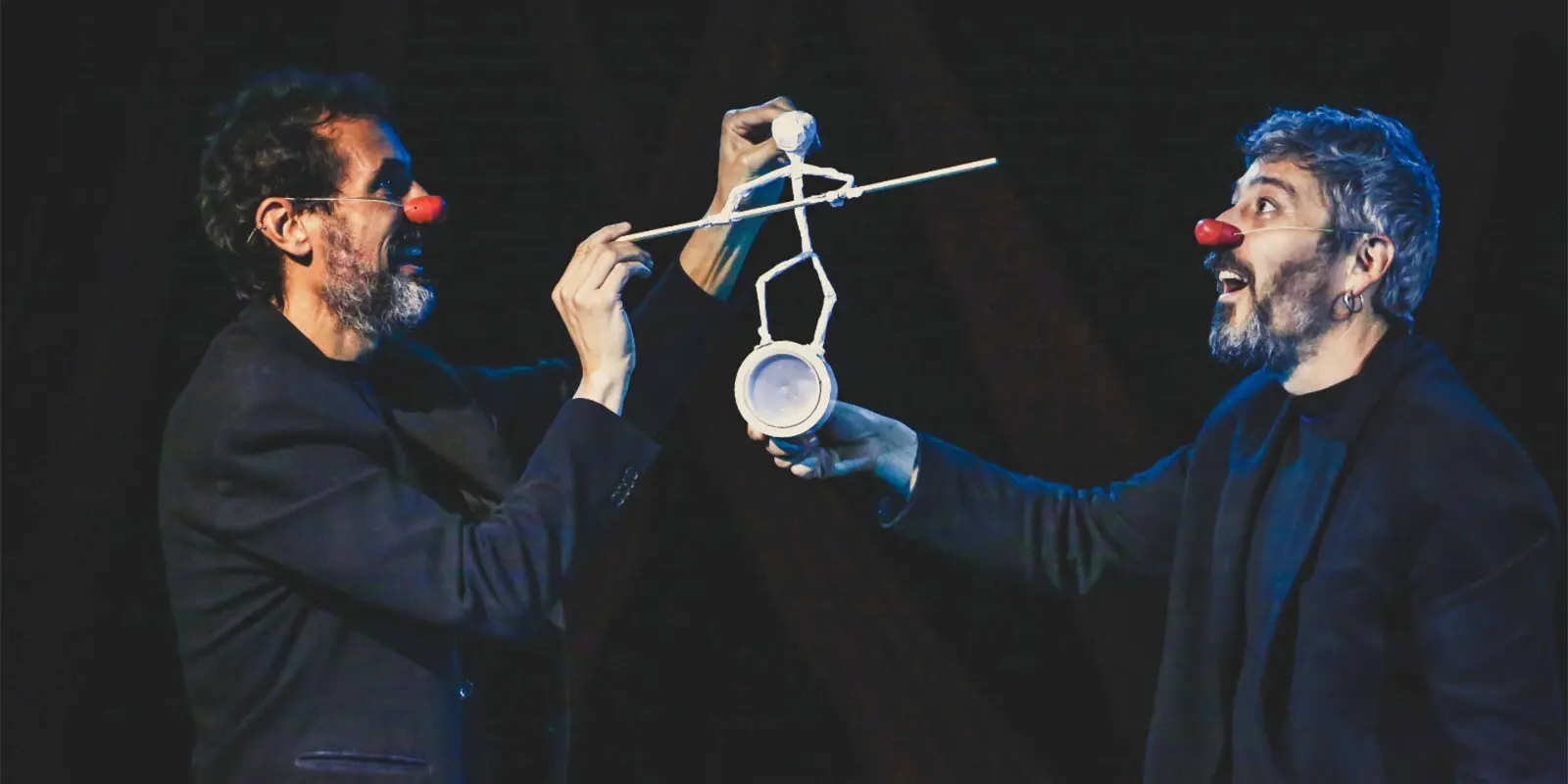 Zwei Männer mit roten Clownnasen stehen sich auf einer dunklen Bühne gegenüber und lachen sich an. Beide tragen dunkle Kleidung und halten gemeinsam eine kleine, weiße Figur, die aus einfachen Materialien geformt ist und auf einem Becher balanciert. Die Szene wirkt humorvoll und konzentriert zugleich, mit Fokus auf das filigrane Spiel zwischen den Darstellern.