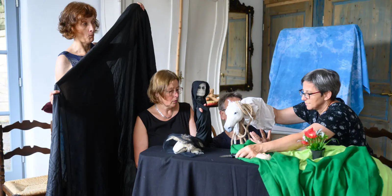 Vier Erwachsene gestalten ein Puppenspiel mit fantasievollen Figuren an einem Tisch, umgeben von Tüchern und Requisiten.