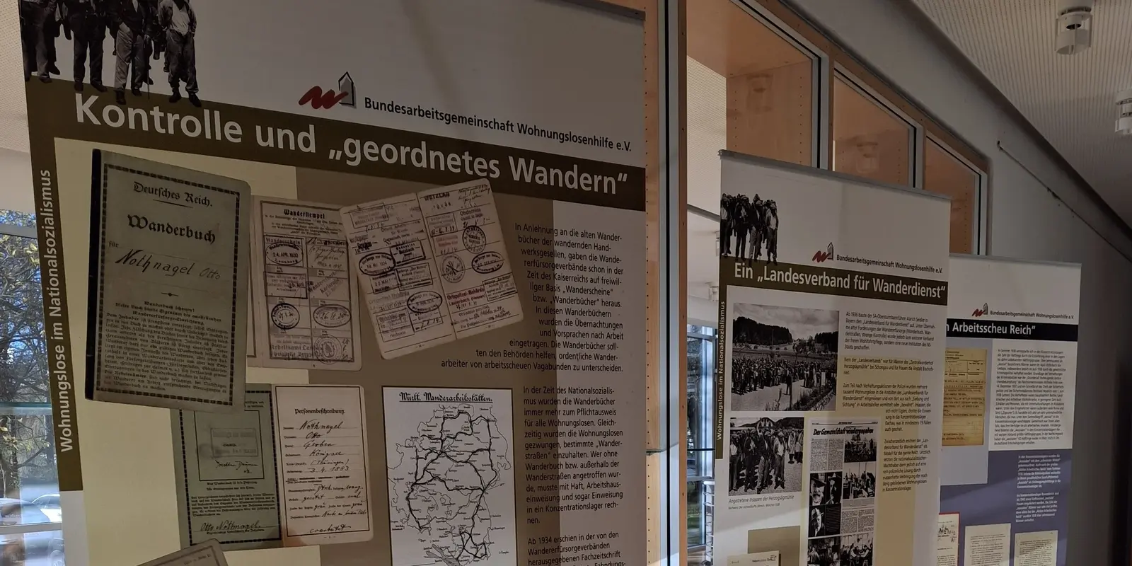 Foto aus der Ausstellung ‚Wohnungslose im Nationalsozialismus‘: Mehrere Roll-up-Tafeln stehen in einem hellen Flur vor großen Fensterflächen. Die vorderen Tafeln tragen Titel wie ‚Kontrolle und „geordnetes Wandern“‘ und zeigen historische Dokumente, Wanderbücher und Karten. Sonnenlicht fällt schräg auf den Holzboden und die Ausstellungswände.