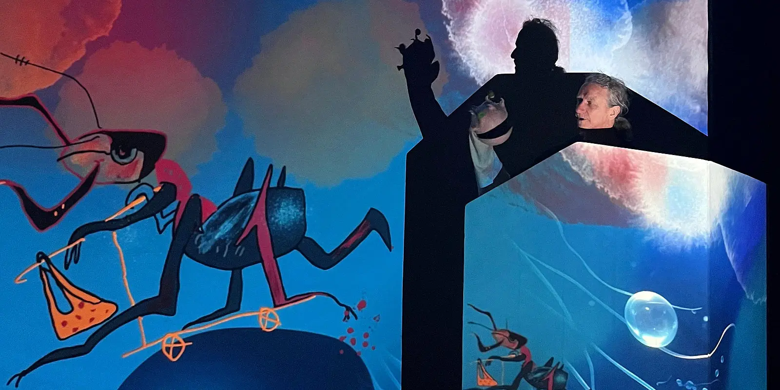 Ein Performer steht als dunkle Silhouette vor zwei leuchtenden Leinwänden mit farbenfrohen animierten Illustrationen – links fantasievolle Vogelwesen, rechts eine blaue Unterwasserwelt mit Ameisen und einer Seifenblase. In der Hand hält er eine kleine Figur.
