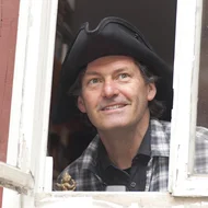 Christoph Buchfink blickt aus einem Fenster mit altem weißem Holzrahmen, er trägt einen dreieckigen Hut.