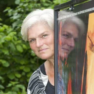 Hiltrud Esther Menz schaut lächelnd hinter einem Plakat hervor, auf dem sich ihr Gesicht spiegelt