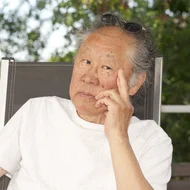 Tadashi Endo sitz in einem Gartenstuhl und stützt seinen Kopf nachdenklich auf seine linke Hand