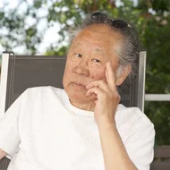 Tadashi Endo sitz in einem Gartenstuhl und stützt seinen Kopf nachdenklich auf seine linke Hand