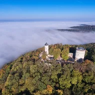 Luftaufnahme der Burg Plesse, umgeben von Wald. Ein Wolkendecke, niedriger als Burg, bedeckt die umgebende Landschaft.