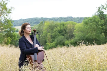 Lucile Chaubard steht mit einem Cello in einem Rapsfeld, im Hintergrund erstreckt sich ein waldiger Hügel.