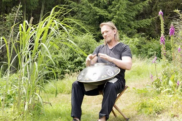 Rolf Predotka (alsia Hangklang) spielt auf einem Stuhl sitzend auf einer Hangdrum. Im Hintergrund ist ein grüner Garten zu sehen.