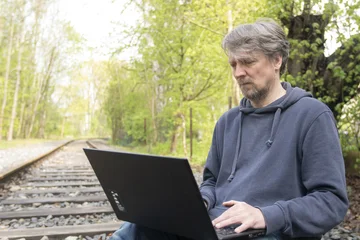 Thomas Kirchberg (alias Tokio) arbeitet auf einer Bahnschiene sitzend an einem Laptop, den er auf seinen Knien stehen hat.