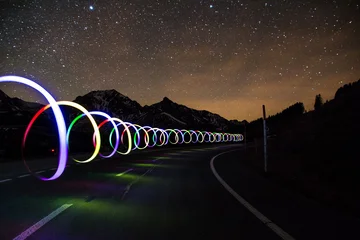 Eine Straße bei nacht mit Sternenhimmel. Über der Straße "schweben" Lichtzeichnungen: eine spiralförmige farbige Lichtröhre