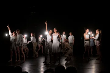 Auf einer abgedunkelten Bühne leuchten kleine weiße Lampen auf Ständern. Schülerinnen des Otto Hahn Gymnasiums sind in Zweierpärchen mit weißen T-Shirt und beigen Hosen oder weisen Kleidern zu sehen. Sie gucken sich gegenseitig in die Augen und halten sich an den Händen. zwei Schülerinnen zeigen mit dem Finger nach oben