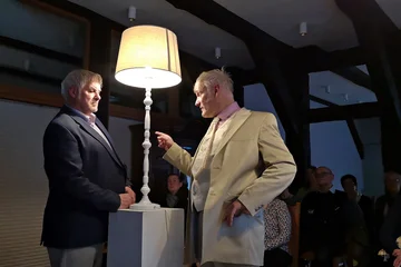Zwei Männer stehen sich in einem dunkel blauen und beigen Anzug gegenüber. Der rechte Mann zeigt auf den linken Mann. Zwischen Ihnen leuchtet eine Stehlampe.
