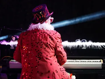 Ein Mann mit einer roten Pailletten-Jacke mit der Aufschrift "ELTON", einer weißen Boafeder und einem roten Paillettenhut sitzt an einem Piano