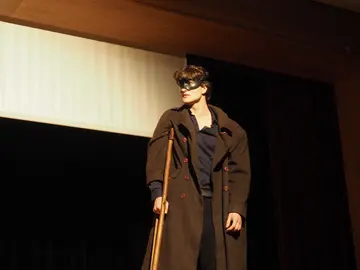 Ein Mann mit Maske / Augenbinde, er trägt eine Art Militärmantel und hstceinecaltmidische Gehhilfe.