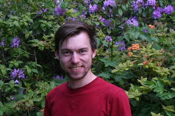 Lukas Keller lächelt mit rotem T-Shirt vor einer Blumenhecke in die Kamera