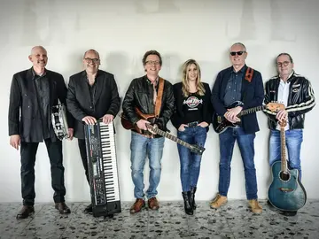 Die Band Rocktail mit 6 Mitgliedern, davon 5 Männer und eine Frau stehen mit ihren Instrumenten in Reihe. Sie tragen alle Jeans und schwarze Jacken.