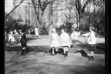 Schwarz-weiß Aufnahme von spielenden Kindern in einem Park im Jahr 1919. Im Vordergrund schwingen zwei Mädchen ein Seil, über das zwei weitere Mädchen springen.