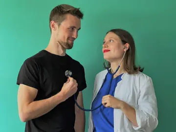 Ein junger Mann in schwarzem T-Shirt und eine junge FRau, die der Kleidung nach Ärztin zus ein scheint (blauer Kasak, weißer Kittel) schauen sich verliebt in die Ohren. Sie hat ihr Stethoskop in den Ohren, das andere Ende presst er auf sein herz.