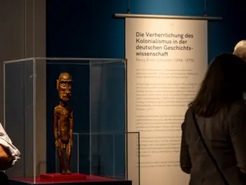Foto aus der Ausstellung: Göttingen kolonial. Im Vordergrund eine moai tangata (= Holzfigur von Rapa Nui/Osterinseln), im Hintergrund eine Textfahne mit der Überschrift Die Verherrlichung des Kolonialismus in der deutschen Geschichtswissenschaft