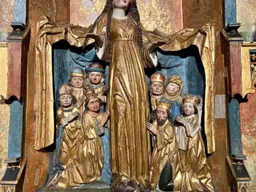 Holzskulptur einer Maria, die ihren Mantel mit blauem Innenfutter ausbreitet und darunter knienden Menschen Schutz bietet