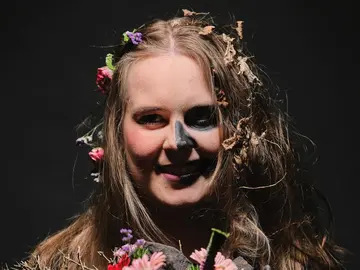 Junge Frau mit halbseitig bemaltem Gesicht, die mit welken Blumen geschmückt ist.