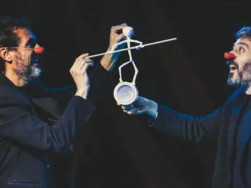 Zwei Männer mit roten Clownnasen stehen sich auf einer dunklen Bühne gegenüber und lachen sich an. Beide tragen dunkle Kleidung und halten gemeinsam eine kleine, weiße Figur, die aus einfachen Materialien geformt ist und auf einem Becher balanciert. Die Szene wirkt humorvoll und konzentriert zugleich, mit Fokus auf das filigrane Spiel zwischen den Darstellern.