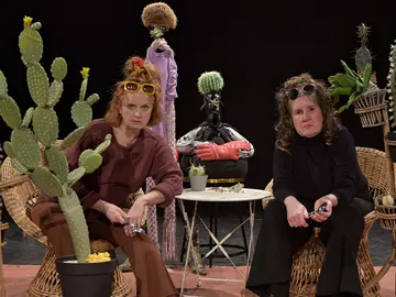Zwei Frauen sitzen in Korbstühlen auf einer Theaterbühne, umgeben von zahlreichen Kakteen in Töpfen und Pflanzenständern. Beide tragen Sonnenbrillen im Haar und halten kleine metallische Gegenstände in den Händen. Im Hintergrund steht eine skurrile Figur mit einem Kaktus als Kopf und roten Boxhandschuhen. Die Szene wirkt humorvoll und absurd, wie ein Teil einer Theaterperformance.
